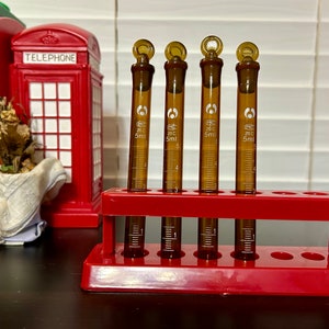 May include: Four amber-colored graduated cylinders, each with a 5ml capacity, are held in a red plastic test tube rack. The cylinders have a white label with text. A red telephone booth figurine is in the background.