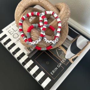 Peut inclure: Trois bracelets de perles rouges, blanches et grises, épelant "OHIO STATE". Les bracelets sont empilés sur un nœud décoratif en bois, posés sur un livre avec des rayures noires et blanches et le texte "NEW YORK".