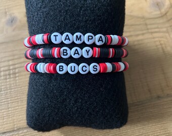 Puede incluir: Tres pulseras de cuentas con las palabras "Tampa Bay Bucs" escritas en letras negras sobre cuentas blancas. Las pulseras son rojas, blancas y negras.