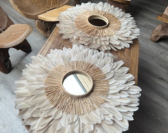 May include: Two circular wall mirrors featuring a central glass pane surrounded by a ring of light tan raffia fibres and an outer layer of layered, off-white feathers. The mirrors are arranged in a sunburst pattern with a soft, textured appearance.