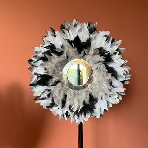 Peut inclure: Un miroir décoratif avec un cadre circulaire, entouré d'un halo de plumes noires, blanches et grises. Le miroir est monté sur un long pied en métal noir, créant une pièce de décoration intérieure unique.