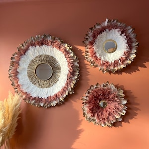 Op de afbeelding: Drie decoratieve wandspiegels met veermotieven. De spiegels hebben een cirkelvormig ontwerp met een centrale spiegel omgeven door witte, roze en goudkleurige veren. Ze zijn gemonteerd op een perzikroze muur.