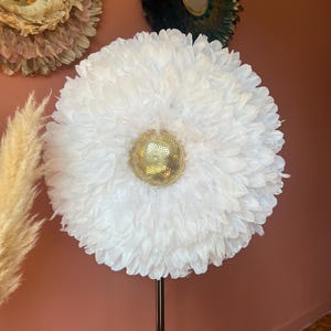 Peut inclure: Une grande décoration murale ronde en plumes blanches avec un centre doré à paillettes, montée sur un support en métal foncé. Deux autres décorations murales en plumes sont visibles en arrière-plan, une verte et une beige.