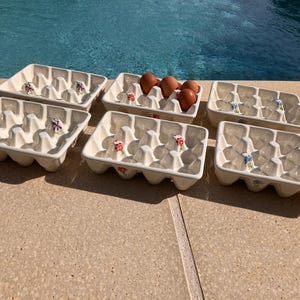 Könnte beinhalten: Sechs cremefarbene Eierhalter aus Keramik, jeweils mit Blumendesign und Platz für sechs Eier. Ein Halter enthält braune Eier. Die Halter sind auf einer Steinoberfläche in der Nähe eines Pools angeordnet.