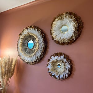 Puede incluir: Tres espejos de pared decorativos con diseños de plumas y espejos. Los espejos son circulares, con un espejo central rodeado de capas de plumas blancas y marrones. Están montados en una pared rosa, creando una estética natural y bohemia.