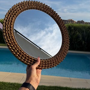 May include: A circular wall mirror featuring a decorative frame covered in numerous small, metallic bronze-coloured shells arranged in a dense, uniform pattern. The reflective glass surface shows a clear reflection of the blue sky and clouds.