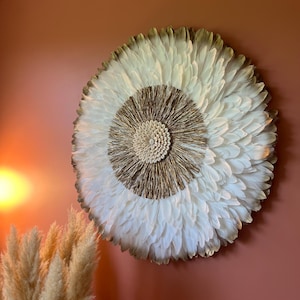 Puede incluir: Decoración mural redonda con un diseño central de fibras naturales tejidas, rodeado de plumas blancas. Los bordes exteriores de las plumas tienen un sutil tono marrón. La pared está pintada de un color marrón rojizo apagado.