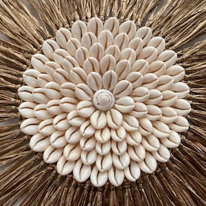 Op de afbeelding: Ronde wanddecoratie met een centraal bloemmotief van witte schelpen. De schelpen zijn omgeven door een rand van gedroogde, goudbruine palmbladeren, wat een natuurlijke, getextureerde uitstraling creëert.
