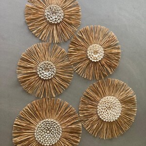Peut inclure: Cinq décorations murales circulaires avec une texture naturelle et tissée. Chaque pièce présente un groupe central de coquillages blancs entouré de fibres brun clair rayonnantes, ressemblant à des fleurs stylisées. Le fond est gris neutre uni.