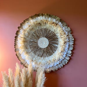 Peut inclure: Décoration murale circulaire en plumes blanches, beiges et marron. Le centre présente un motif texturé, monté sur un mur rose. Des herbes de la pampa sont au premier plan, ajoutant à l'esthétique naturelle.