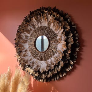Peut inclure: Un miroir mural rond avec un design décoratif en plumes. Le miroir est entouré de couches de plumes marron, blanches et noires, créant un cadre circulaire texturé. Le miroir lui-même est petit et rond, reflétant l'environnement.