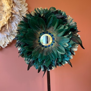 Peut inclure: Décoration murale circulaire composée de plumes vertes et noires, avec un petit miroir rond au centre. Le miroir est entouré d'accents de plumes de paon. Une autre décoration murale similaire est visible en arrière-plan.