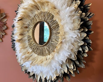 May include: A circular mirror with a gold frame, encircled by a halo of white, gold, and black feathers. The mirror is mounted on a dark metal stand, creating a striking wall art piece.