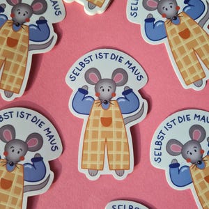 Könnte beinhalten: Aufkleber mit einer Comic-Maus, die ihre Armmuskeln anspannt und ein blaues Hemd und eine gelbe Latzhose trägt. Der Text "SELBST IST DIE MAUS" ist auf jedem Aufkleber aufgedruckt. Auf rosa Hintergrund.