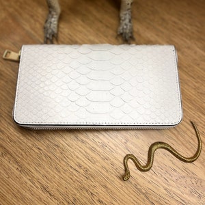 May include: A rectangular white wallet with a textured snakeskin pattern and a gold-toned metal zipper pull. The wallet features a structured design with visible stitching along the edges and a smooth, uniform finish across the front surface.