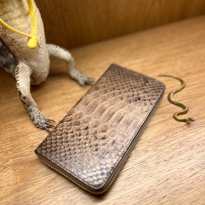 May include: A rectangular wallet featuring a textured, brown and tan snakeskin pattern with a metallic zipper closure along the edge. The wallet has a distinct scale-like surface texture and is positioned next to a lizard figurine and a small snake.