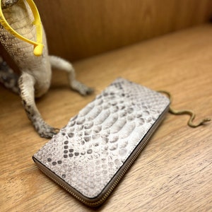 May include: A rectangular wallet featuring a textured, light gray and white snakeskin pattern with a metallic zipper closure running along the perimeter. The wallet has a distinct scale-like surface texture and is positioned at an angle.