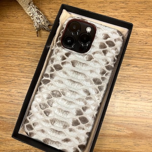 Op de afbeelding: Een smartphonehoesje met een gestructureerd slangenleerpatroon in gebroken wit en grijs. Het hoesje is rechthoekig met afgeronde hoeken en bevat een uitsparing voor een camerasysteem met drie lenzen. Het wordt gepresenteerd in een zwarte doos.