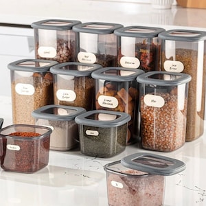 May include: A set of 12 clear plastic storage containers with grey lids. The containers are filled with various dry goods, such as rice, pasta, and spices. Each container has a label with a handwritten name, such as "Rice", "Pasta", and "Spices". The containers are arranged in a pyramid shape on a white worktop. 12PCS SET
