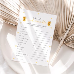 Può includere: Una carta di gioco per la festa di addio al nubilato stampabile con il titolo "Bridal Word Scramble". La carta contiene un elenco di parole relative al matrimonio da decifrare. La carta è decorata con piccoli cuori gialli e illustrazioni di barattoli di miele.