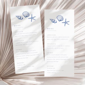 May include: Two white menu cards with a blue seashell and starfish design. The menu includes appetizers, antipasta, entree, and dessert options. The cards are displayed on a light-colored, textured background.