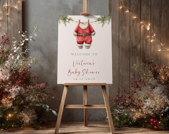 May include: A baby shower sign on a wooden easel. The sign shows a watercolour illustration of a red Santa suit hanging on a washing line. Text reads "Welcome to Victoria's Baby Shower" and the date 14.12.2025.
