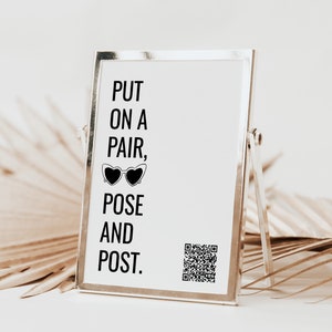 May include: A silver picture frame with a white background and black text that reads "PUT ON A PAIR, POSE AND POST."  A pair of sunglasses and a QR code are also visible on the frame.