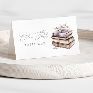 May include: A white place card with the text "Chloe Todd" and "TABLE ONE" in elegant script. A watercolor illustration of stacked books with flowers adorns the card. The card is on a wooden tray.