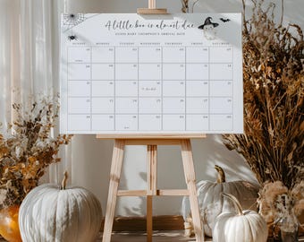 May include: A white calendar on a wooden easel with the text "A little boo is almost due." The calendar has a grid layout with dates and is decorated with Halloween-themed graphics. Pumpkins and dried plants are in the foreground.