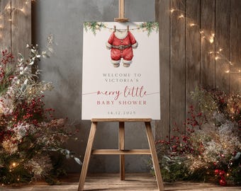 May include: A wooden easel holds a sign for a baby shower. The sign features a watercolour illustration of a Santa Claus outfit hanging on a clothesline. Text reads "Welcome to Victoria's Merry Little Baby Shower" and the date.
