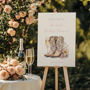 Peut inclure: Une pancarte blanche avec le texte "Bienvenue au Bridal Shower d'Amelia" en écriture rose. La pancarte présente également une illustration aquarelle de deux bottes de cowboy, une coupe de champagne et des fleurs sauvages. La date "08.02.202X" est imprimée en bas de la pancarte.