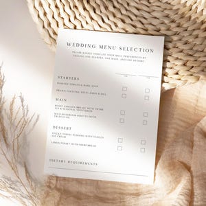 Op de afbeelding: Een witte trouwmenukaart met zwarte tekst, met een lijst van voorgerechten, hoofdgerechten en desserts. De kaart ligt op een beige stoffen ondergrond, met een gevlochten mand en gedroogde planten op de achtergrond.