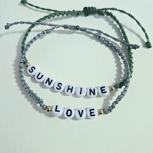 May include: Two macrame bracelets. One is green, the other is gray. The gray bracelet has white letter beads that spell "SUNSHINE" and "LOVE". The bracelets have adjustable sliding knots.