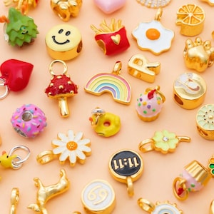 May include: A collection of gold-toned charms with various designs, including a smiley face, rainbow, mushroom, and a fried egg. Other charms feature a donut, flower, and a black charm with the numbers 11:11. The charms are on a light peach background.