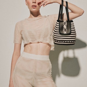May include: A woman wearing a sheer, white crop top and matching pants holds a black and white striped tote bag with a black leather flap.