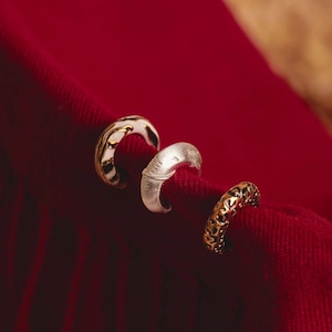 May include: Three ear cuffs in gold, silver, and gold-tone metal rest on a deep red fabric. The cuffs have unique designs: one with a white enamel pattern, one with a textured surface, and one with an openwork design.