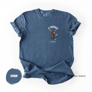 Puede incluir: Camiseta azul denim con mangas enrolladas. La camiseta presenta un gráfico de un toro con traje y portátil, con el texto "IT WORKED" y "ON MY ASSUMPTION".