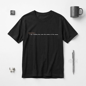 Peut inclure: T-shirt noir avec le texte "* Wrangling... L Tip: Herding divs into the center of the screen" en police simple sans empattement. Le t-shirt est présenté sur une surface blanche, avec une tasse grise, un stylo et une figurine de chat.