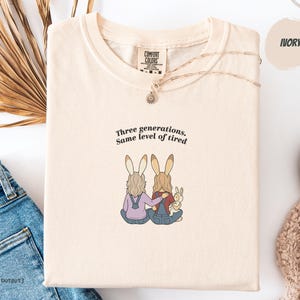 Peut inclure: T-shirt ivoire avec l'inscription "Three generations. Same level of tired" et un dessin de deux lapins en salopette. Un collier doré est posé sur le t-shirt. Marque Comfort Colors.