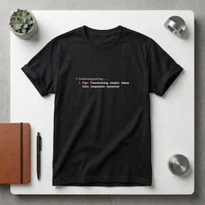 Op de afbeelding: Zwart T-shirt met opgerolde mouwen. Het shirt heeft de tekst "* Gobbledygooking... L Tip: Translating simple ideas into corporate nonsense" in oranje en wit. Het shirt wordt gepresenteerd op een wit oppervlak.