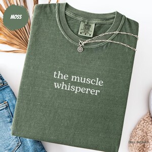 May include: A moss green t-shirt with the phrase "the muscle whisperer" printed in white. The shirt is folded and has a silver necklace with a sun pendant. The tag reads "Comfort Colors".