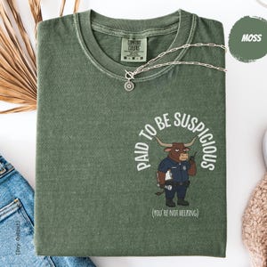 Könnte beinhalten: Ein moosgrünes T-Shirt mit Rundhalsausschnitt und einer Grafik eines braunen Stiers als Polizist, der ein Klemmbrett und eine Taschenlampe hält. Der Text lautet PAID TO BE SUSPICIOUS und (YOU'RE NOT HELPING). Das Shirt hat ein Comfort Colors Etikett.