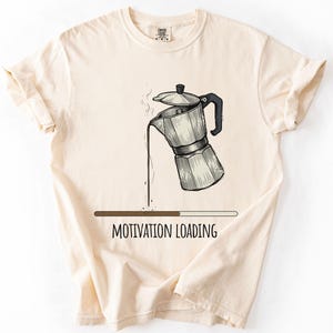 May include: Cream-colored t-shirt featuring a coffee pot graphic with coffee pouring out. Below the graphic is a loading bar with the text "MOTIVATION LOADING". The shirt has short sleeves and a crew neck.