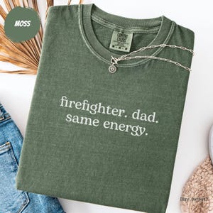 May include: A moss green t-shirt with the text "firefighter. dad. same energy." in white. The shirt has a crew neck and short sleeves. A silver necklace with a sun pendant is on the shirt. The shirt is folded on a white surface.