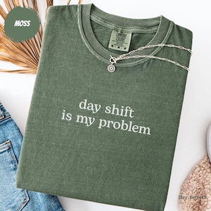 Könnte beinhalten: Ein moosgrünes T-Shirt mit dem weißen Aufdruck "day shift is my problem". Eine silberne Halskette mit Sonnenanhänger liegt auf dem Shirt. Das Shirt ist gefaltet und wird mit einer Jeans präsentiert.