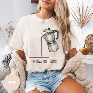 以下が含まれることがあります： アイボリー色のTシャツ。コーヒーポットのグラフィックと「MOTIVATION LOADING」の文字がプリントされています。グラフィックは、ポットからコーヒーが注がれている様子を描いています。Tシャツはデニムショーツとベージュのカーディガンと組み合わせています。