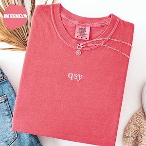 May include: A watermelon-coloured short-sleeve t-shirt with a crew neckline and the text qsy embroidered in white on the centre chest. The shirt features a Comfort Colors label at the inner neck and is paired with a gold-toned chain necklace with a pendant.