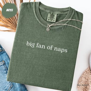 Könnte beinhalten: Ein moosgrünes T-Shirt mit dem weißen Aufdruck "big fan of naps". Das Shirt hat einen Rundhalsausschnitt und kurze Ärmel. Eine silberne Halskette mit Sonnenanhänger liegt über dem Shirt. Das Shirt ist auf einer weißen Oberfläche gefaltet.