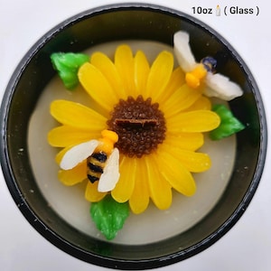 May include: A sunflower-shaped candle with yellow petals and a brown center, featuring two bee-shaped wax figures. The candle is in a black container and includes green leaf accents. The text "10oz (Glass)" is visible.
