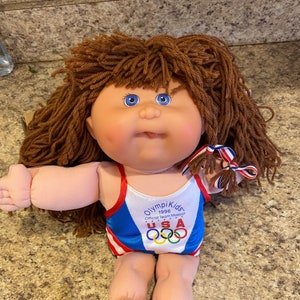May include: A Cabbage Patch Kid doll wearing a blue and red swimsuit with the Olympic Kids 1996 logo and the words "Official Team Mascot" on the front. The doll has brown yarn hair and blue eyes.
